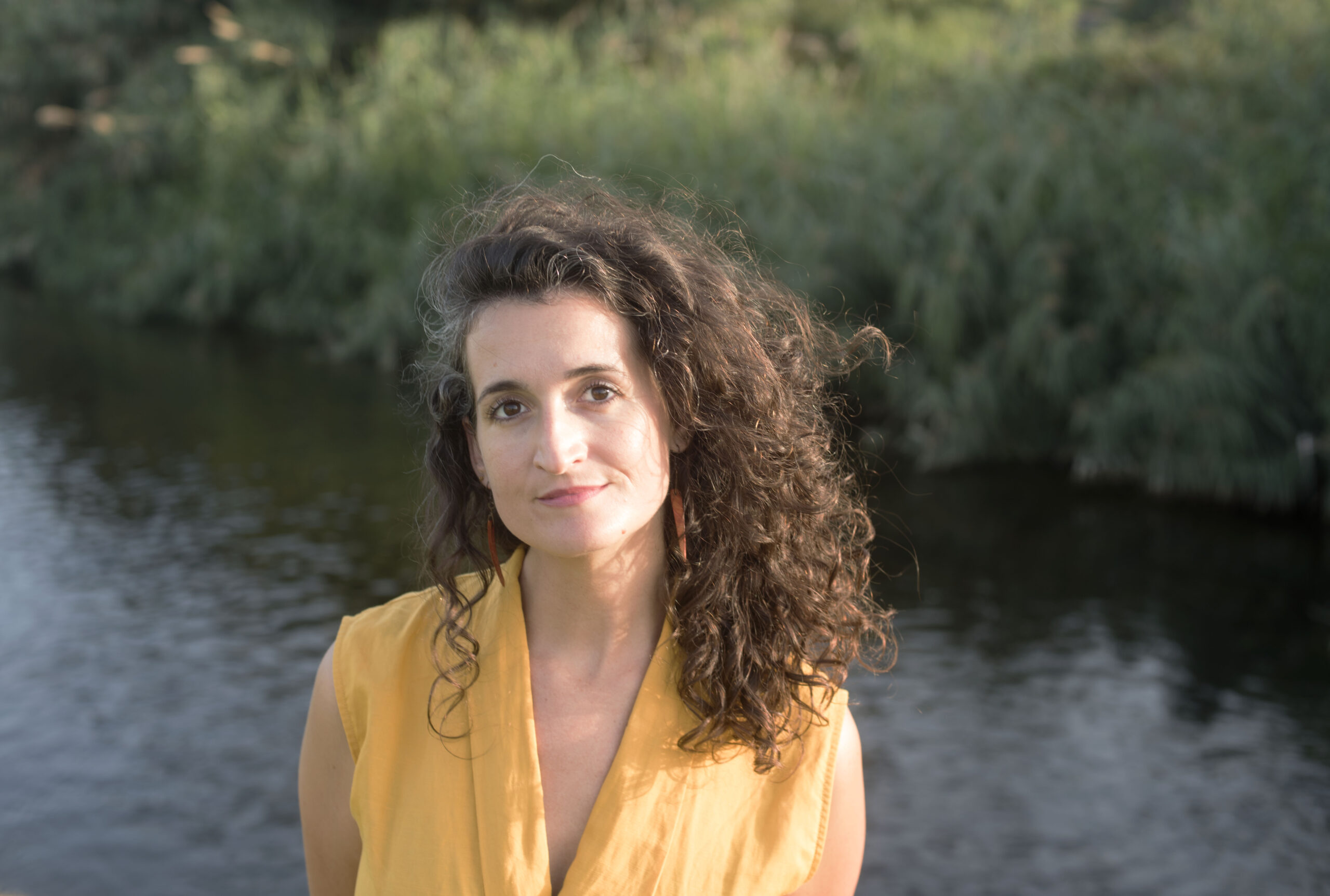 _DSC1953 Portrait of Judit Saez in a yellow top, standing by a lake with reeds in the background