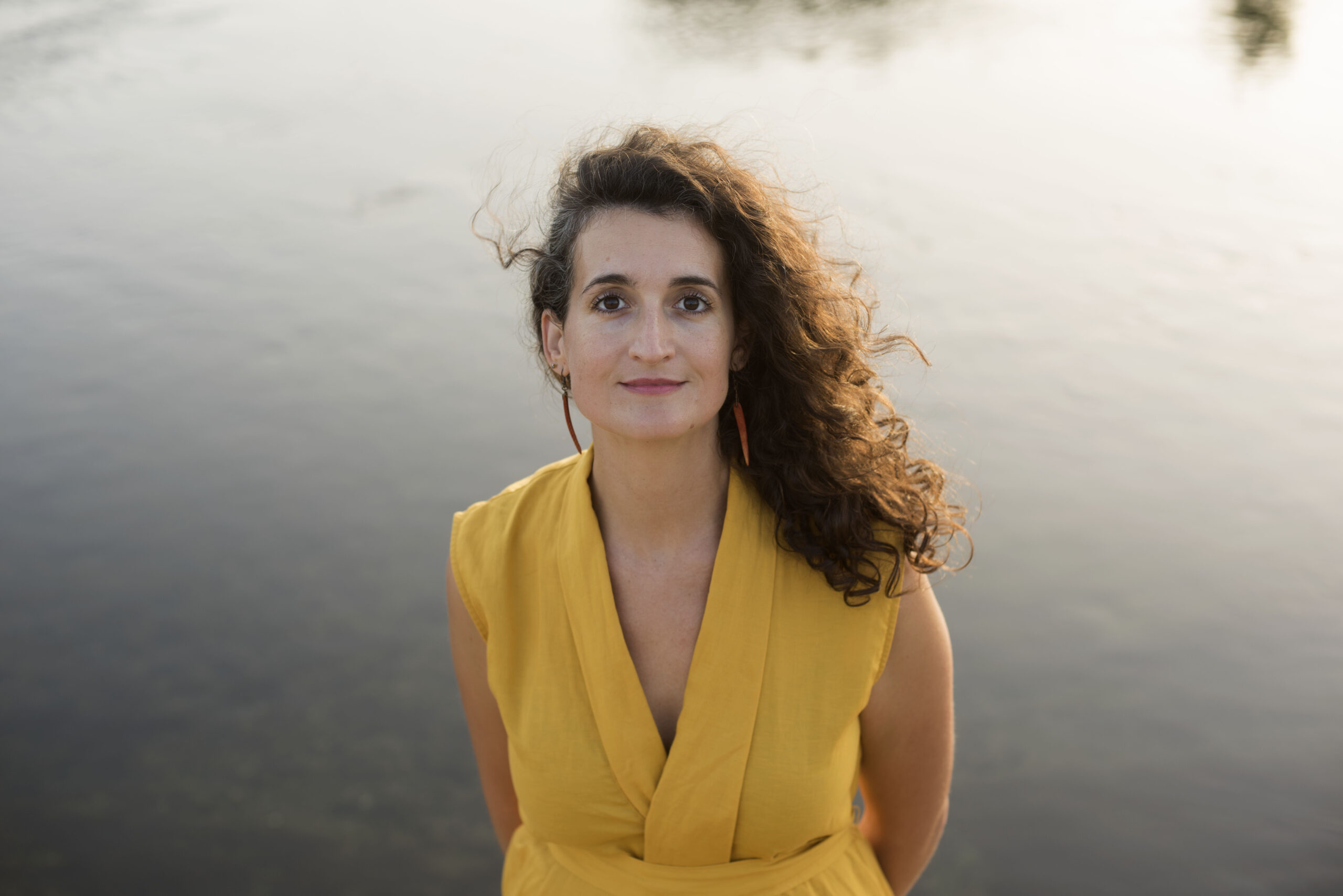 _DSC1917 Judit Saez standing by calm water at golden hour, wearing a yellow top