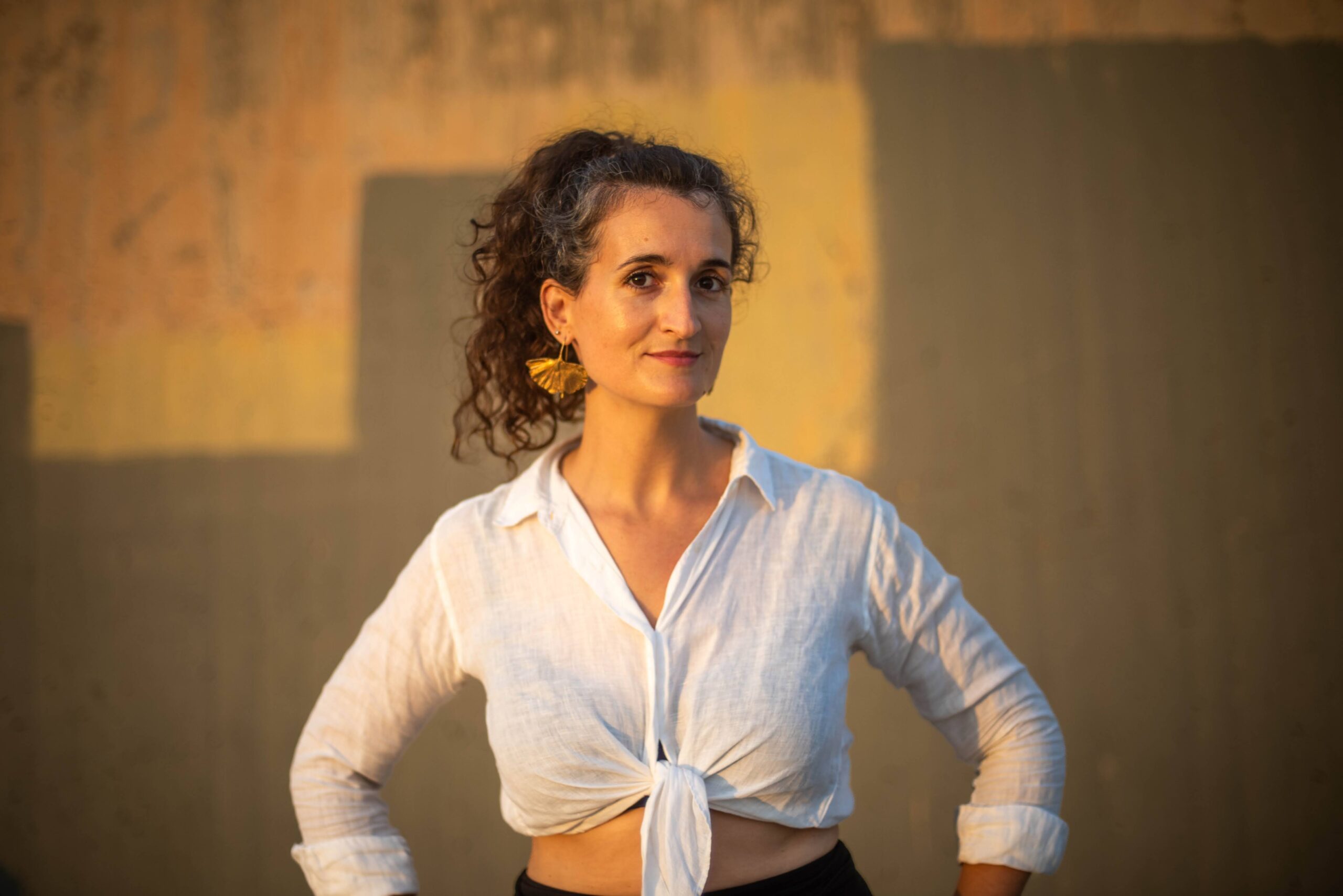 _DSC2483 Confident portrait of Judit Saez in a white blouse, hands on hips, against a warm textured wall